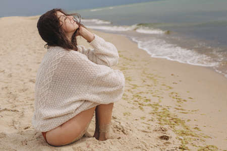 Carefree beautiful woman with windy curly hair sitting on sandy beach at sea, calm moment. Stylish young happy female in knitted sweater relaxing on coast. Summer vacation moodの写真素材