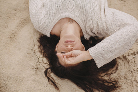 Beautiful woman lying on sandy beach, carefree tranquil moment. View above. Stylish young female in knitted sweater enjoying vacation and relaxing on coastの写真素材