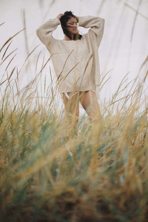 Beautiful stylish woman in knitted sweater and with windy hair posing among wild grass at sea. Authentic carefree moment. Fashionable young female standing on windy coast. Tranquility. Verticalの写真素材