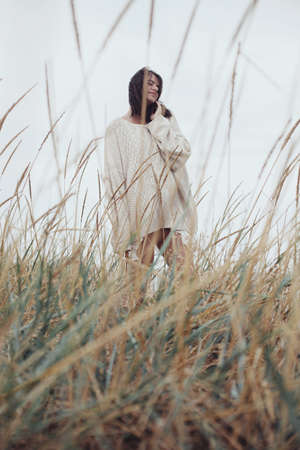 Beautiful stylish woman with windy hair in knitted sweater posing among wild grass. Carefree moment, stylish image. Fashionable young female standing on windy coast. Authentic and Tranquilityの写真素材
