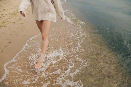 Feet in splashing waves. Carefree beautiful woman running barefoot on sandy beach with cold sea...