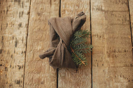 Stylish christmas gift wrapped in fabric with fir branch on rustic wooden table top view. Zero waste winter holidays and gift. Furoshiki eco friendly present. Happy holidays and Merry Christmasの写真素材