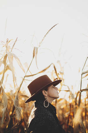 Beautiful stylish woman in brown hat and vintage coat relaxing in autumn maize field in sunset light. Portrait of fashionable young hipster female in retro outfit enjoying evening in autumn cornの写真素材