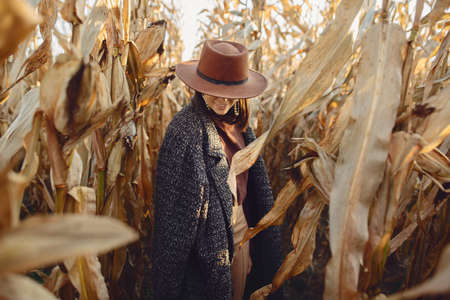 Portrait of happy stylish woman in brown hat and vintage coat walking in autumn maize field in sunset light. Fashionable young hipster female in retro outfit standing in autumnal cornの写真素材