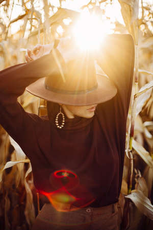 Portrait of stylish woman in hat and brown clothes posing in autumn maize field in sunset sunlight. Fashionable attractive young female  standing in autumnal corn in evening countrysideの写真素材