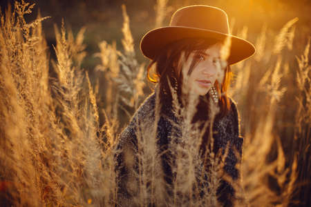Portrait of beautiful stylish woman in hat standing in grass in warm sunset light in autumn field....