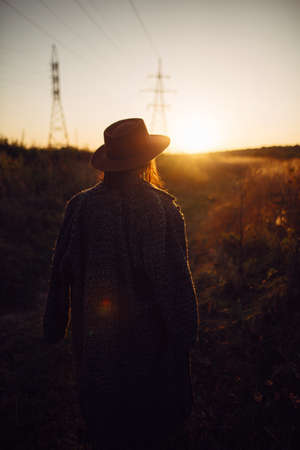Beautiful stylish woman in hat and vintage coat walking in autumn field in warm sunset light. Tranquil atmospheric moment. Fashionable attractive young female enjoying sunset in rural countrysideの写真素材