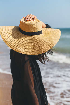 Stylish carefree woman holding hat and relaxing on sunny beach at sea, close up. Summer vacation. Young fashionable female in straw hat and light shirt enjoying holiday on tropical islandの写真素材
