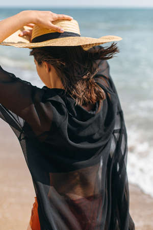 Beautiful stylish woman in hat walking on windy beach at sea waves. Summer vacation. Fit sexy young female in light shirt and straw hat relaxing on tropical islandの写真素材