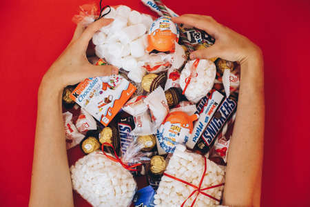 Lviv, Ukraine - December 1, 2017: Hands holding many sweets and candy bars on red background, top view. Assortment of Raffaello, Snickers, Kinder, Ferrero, M&M's, Milka, Raffaello. Halloween candyのeditorial素材