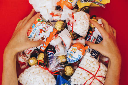 Lviv, Ukraine - December 1, 2017: Hands holding many sweets and candy bars on red background, top...