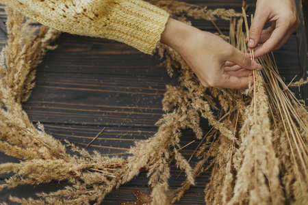 Hands holding dry grass and making stylish autumn boho wreath with wildflowers and herbs on rustic wooden table. Holiday workshop. Florist in yellow sweater making rustic autumn wreathの写真素材