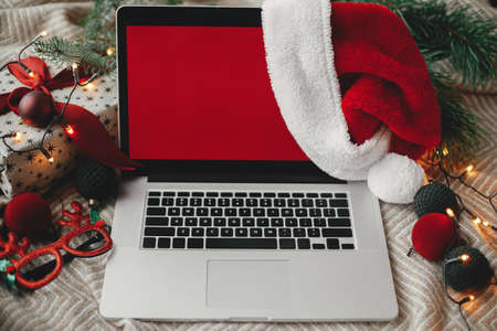 Christmas sales and shopping online. Laptop with red empty screen in santa hat on cozy bed with stylish christmas ornaments, present and festive lights. Space for text. View above. Freelanceの写真素材