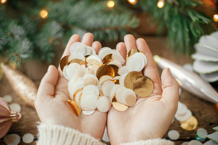 Hands holding golden confetti on background of christmas decorations, baubles and pine branches in lights on rustic wood. Space for text. Happy holidays. Seasons greetingsの写真素材