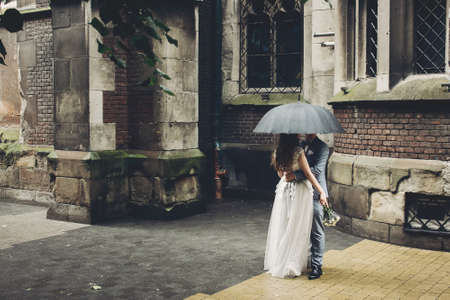 Stylish bride and groom kissing under umbrella on background of old church in rain. Provence wedding. Beautiful wedding couple embracing under black umbrella in rainy street. Romantic momentの写真素材