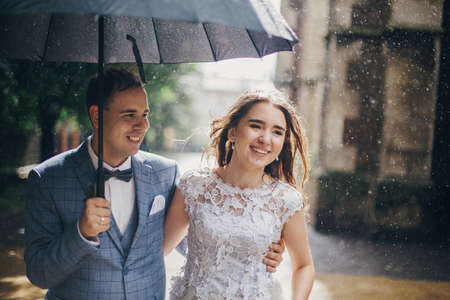 Stylish happy bride and groom walking and smiling on background of old church in rain. Provence wedding. Beautiful wedding couple embracing together under umbrella in sunny rainy street.の写真素材