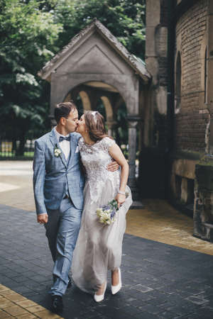 Beautiful emotional wedding couple kissing and walking in european city. Provence wedding. Stylish happy bride and groom walking on background of church in sunny streetの写真素材