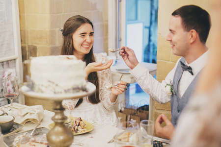 Stylish happy bride and groom tasting delicious modern cake in stylish restaurant. Provence wedding reception. Beautiful wedding couple smiling and eating slice of delicious wedding cakeの写真素材