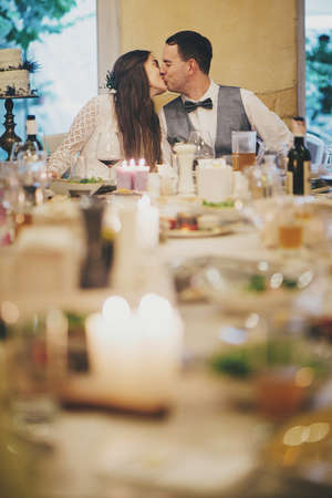Stylish happy bride and groom kissing on background of delicious festive table with food and drinks in restaurant. Provence wedding reception. Beautiful wedding couple celebrating weddingの写真素材