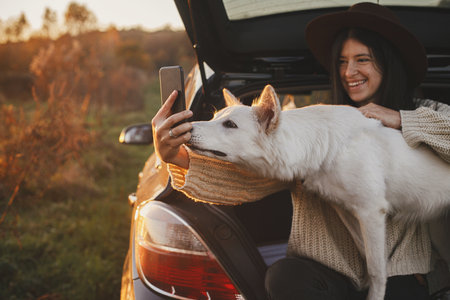 Autumn road trip with pet and travel. Stylish hipster woman taking selfie photo with cute dog in car trunk in sunset light in field. Young female in hat and sweater using phone with sweet white dogの写真素材