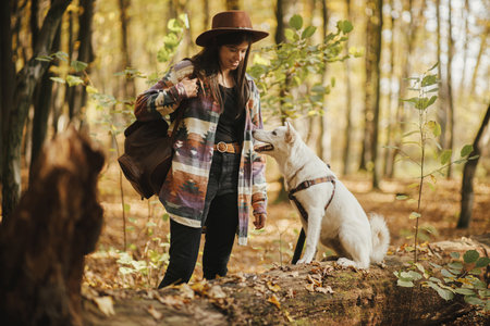 Stylish woman in hat training cute dog sitting on old stump in sunny autumn woods. Young female traveler with swiss shepherd white dog in beautiful forest. Travel and learning with petの写真素材