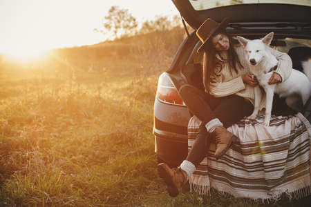 Stylish woman in hat and sweater sitting and hugging cute dog in car trunk in warm sunset light in autumn field. Road trip with pet.Young hipster female travelling with sweet dog. Space for textの写真素材
