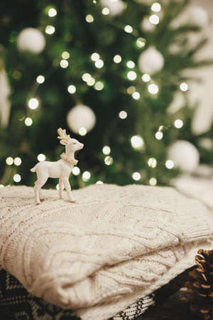 Cozy knitted sweaters with reindeer toy and pine cone on rustic wood on background of christmas tree lights. Pile of stylish sweaters in festive decorated scandinavian room.  Winter holidaysの写真素材