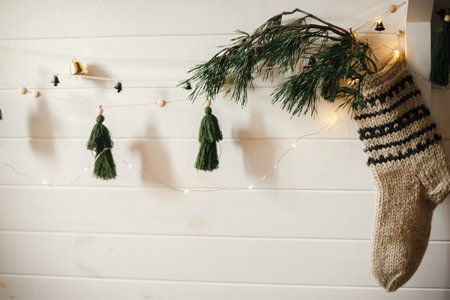 Christmas rustic stocking with fir branch and stylish garland with bells and trees on white wooden wall with christmas lights. Festive decorated atmospheric scandinavian room. Modern handmade decorの写真素材