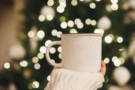 Stylish cup in hand on background of christmas tree in lights in evening room. Cozy winter time. Space for text. Female hand in sweater holding ceramic cup with tea in festive atmospheric roomの写真素材