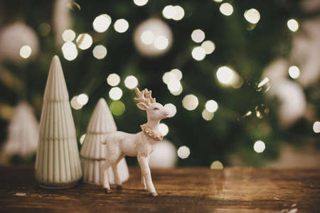 Stylish reindeer toy and christmas trees on rustic wood on background of christmas lights. Beautiful little christmas scene in festive scandinavian room. Space for text. Merry christmasの写真素材