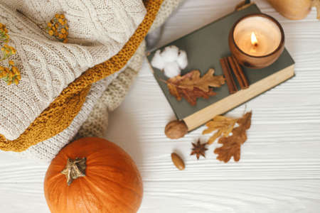 Cozy autumn days, slow living. Pumpkin, cozy sweaters, autumn leaves, burning candle and vintage book on white wooden background in room.Happy Thanksgivingの写真素材