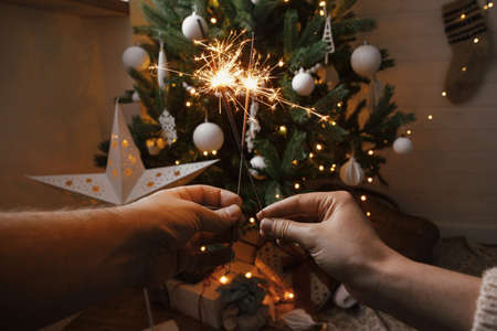 Couple celebrating with firework bengal lights on background of christmas tree and glowing star. Happy New Year! Hands holding burning sparklers in festive scandinavian room. Atmospheric momentの写真素材