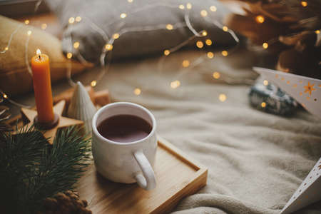 Warm cup of tea with christmas lights, stars, pine trees, candle and pillows on soft bed. Cozy home. Winter hygge. Festive holiday decorations in scandinavian evening room. Atmospheric  momentの写真素材