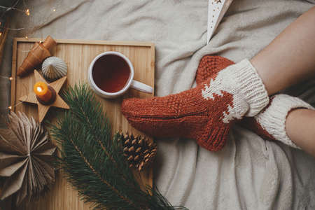 Woman feet in woolen socks on soft bed with warm cup of tea, christmas stars, golden lights, trees, candle and pillows. Top view. Cozy moments at home. Girl relaxing in scandinavian evening roomの写真素材