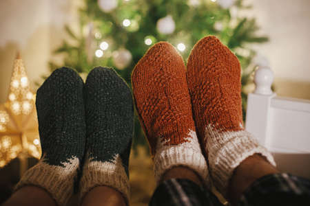 Couples feet in cozy woolen socks on background of christmas tree in lights in festive evening room. Celebrating winter holidays together, cozy family moments at home. Stylish warm socksの写真素材