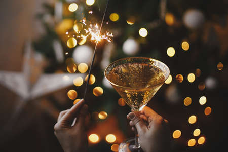 Happy New Year! Hands holding burning sparkler and champagne glass on background of christmas tree lights and star. Firework bengal light and drink in woman hands. Atmospheric momentの写真素材