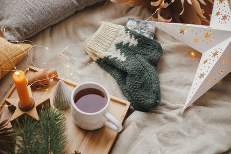 Warm cup of tea, woolen socks, christmas stars, golden lights, trees, candle and pillows on soft...