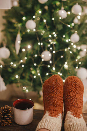 Cozy winter moments at home. Woman feet in cozy woolen socks and cup of warm tea on background of christmas tree in lights in festive evening room. Stylish warm socks on female legsの写真素材