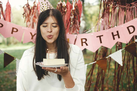 Stylish happy woman in party hat blowing candle on piece of birthday cake and making a wish on background of pink happy birthday garland in park. Celebrating birthday at picnic party outdoor.の写真素材