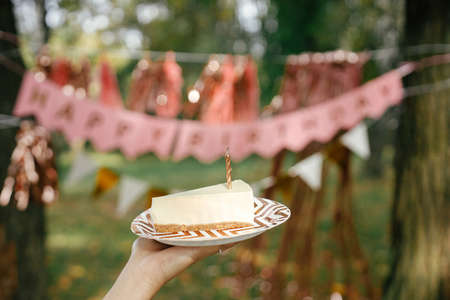 Piece of modern birthday cake with candle in woman hand on background of pink happy birthday garland...
