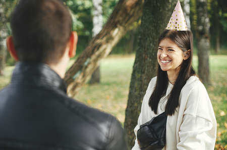 Stylish young happy woman in party hat celebrating birthday with family and friends at picnic party outdoor. Man congratulating female friend on her birthday. Family gathering in parkの写真素材