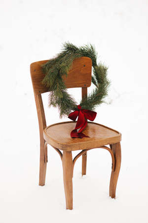 Merry Christmas! Christmas wreath on rustic chair in snowy winter field. Winter holidays in countryside. Stylish xmas wreath with pine branches and red bow on wooden chair. Atmospheric timeの写真素材