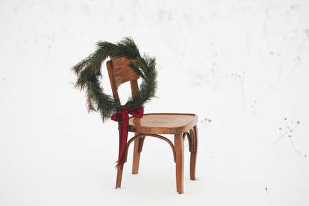 Christmas wreath on rustic chair in snowy winter field. Merry Christmas! Winter holidays in countryside. Space for text. Stylish xmas wreath with pine branches and red bow hanging on wooden chairの写真素材