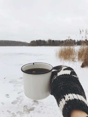 Hand in cozy glove holding warm cup of tea on background of snowy lake in winter. Hiking and traveling in cold winter season. Warm drink. Space for textの写真素材