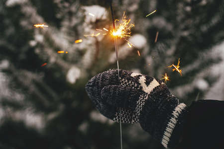 Hand in cozy glove holding burning sparkler on background of pine tree branches in snow. Happy New Year! Atmospheric magic moment. Woman hand with glowing firework in evening. Happy Holidays!の写真素材