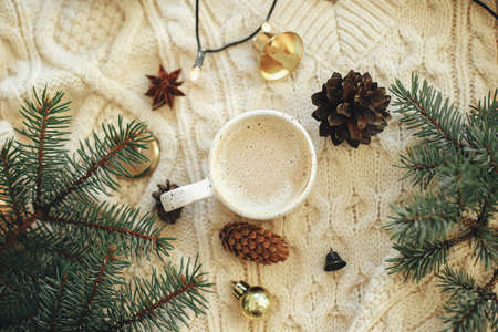 Warm coffee in stylish cup, anise star, fir branches, ornaments, pine cones and warm lights on cozy knitted background. Top view. Atmospheric christmas time and hygge winter homeの写真素材