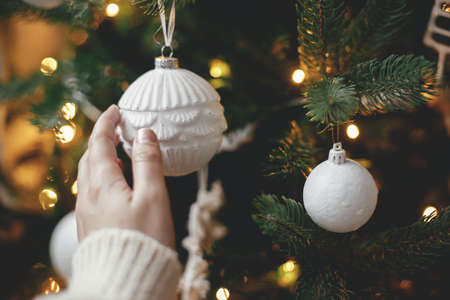 Hand in cozy sweater holding modern white bauble at christmas tree lights. Woman decorating stylish christmas tree with vintage ornament. Winter holidays preparations. Atmospheric joyful momentsの写真素材