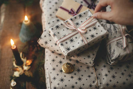 Hand opening stylish christmas gift wrapped in paper on rustic wooden table with candles, bells, fir...