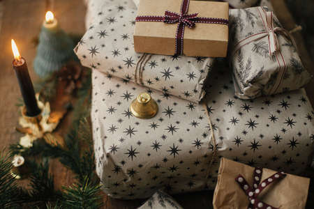 Merry Christmas! Stylish christmas gifts wrapped in craft paper, vintage candles, fir branches and bells on rustic wood. Stylish scandinavian xmas presents, atmospheric winter time in countrysideの写真素材