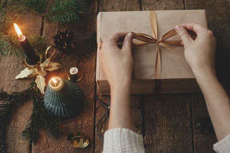 Hands wrapping stylish christmas gift in craft paper on rustic wooden background with candle, scissors, fir branches. Modern simple eco friendly xmas present, scandinavian atmospheric moody imageの写真素材
